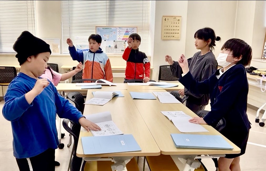 机を囲んで手を挙げながら英語を練習する子どもたち